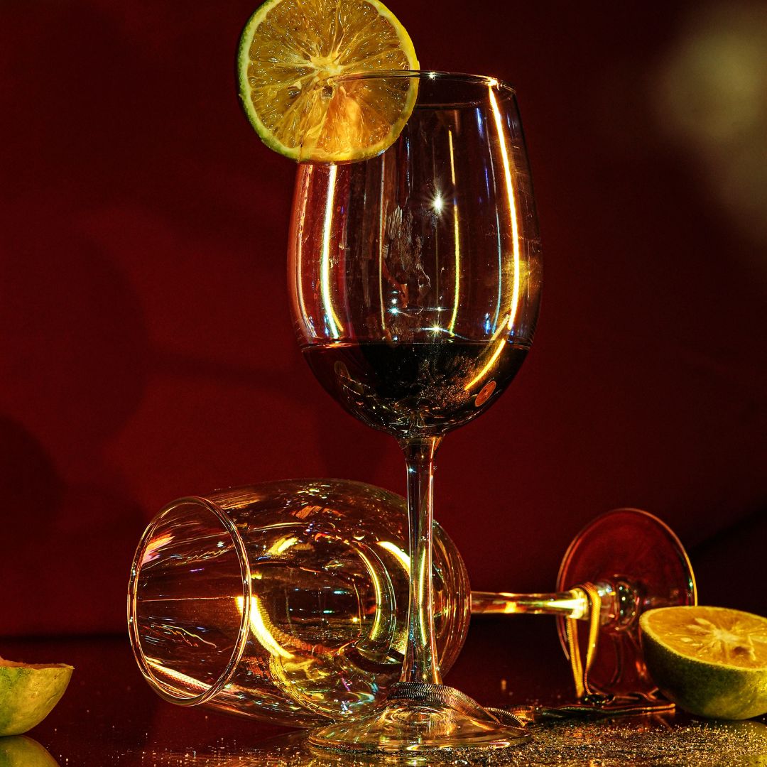A set of rainbow-colored long stem wine glasses with one glass upright displaying red wine, surrounded by two other glasses that are turned on their sides. The scene includes lemon slices and a reflective surface.