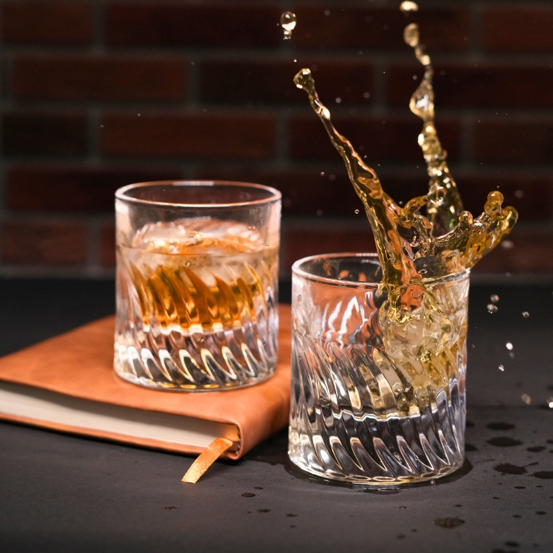 Two glasses with liquid, one being splashed against a brick wall background.