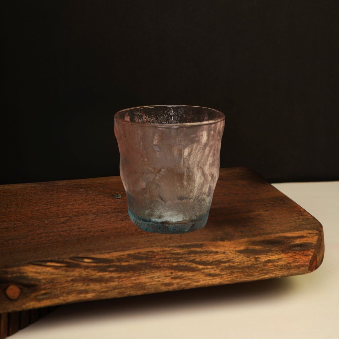 A single glass tumbler with a glacier-textured surface and an ombre fade from pink to blue, placed on a wooden board.